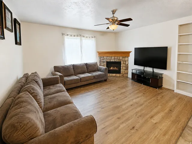 Spacious living area featuring wood-finish flooring, a corner stone-faced fireplace with a wood mantle, and a ceiling fan with integrated lighting