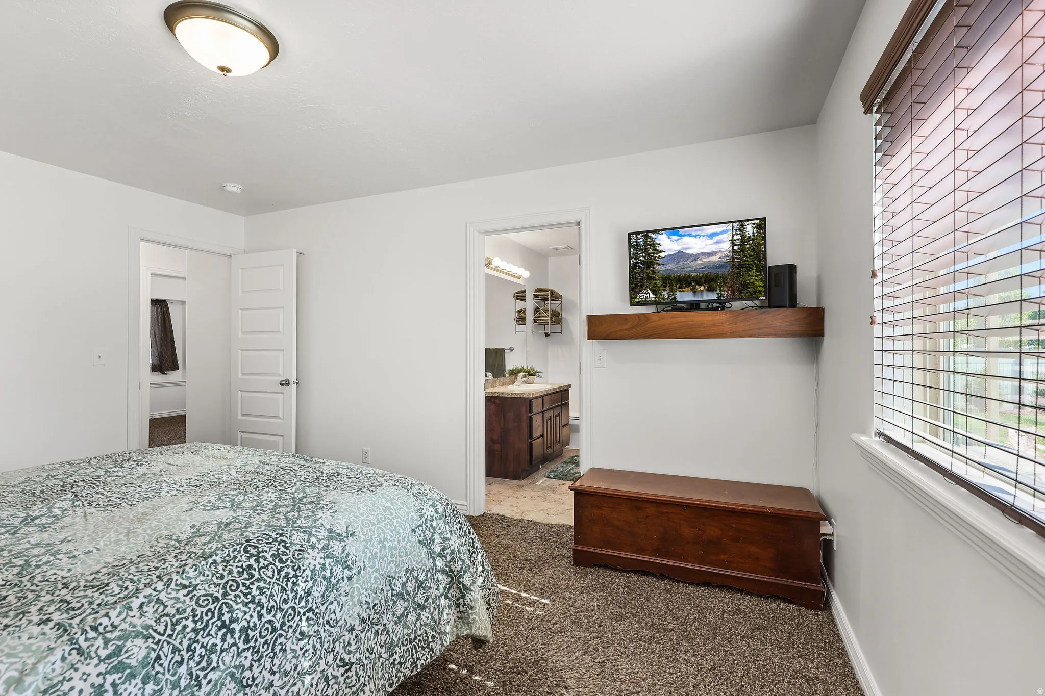 Carpeted bedroom featuring a window with blinds, a wood-finish floating shelf, a ceiling-mounted light fixture, and white trim