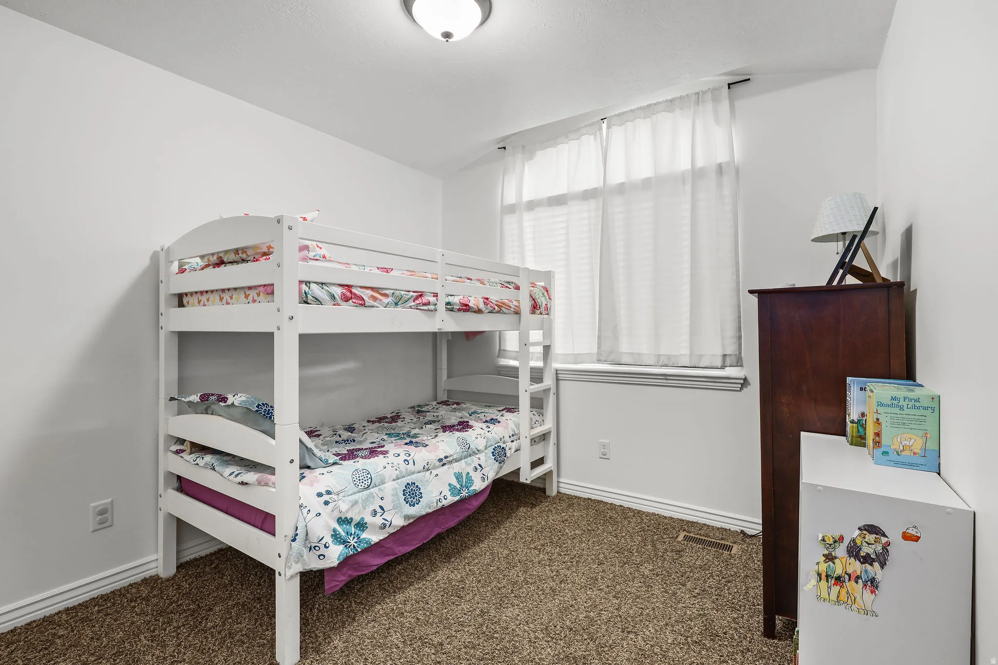 Neutral-toned carpeting grounds this room featuring a window with blinds and sheer white curtains, white baseboards, and a flush-mount ceiling light