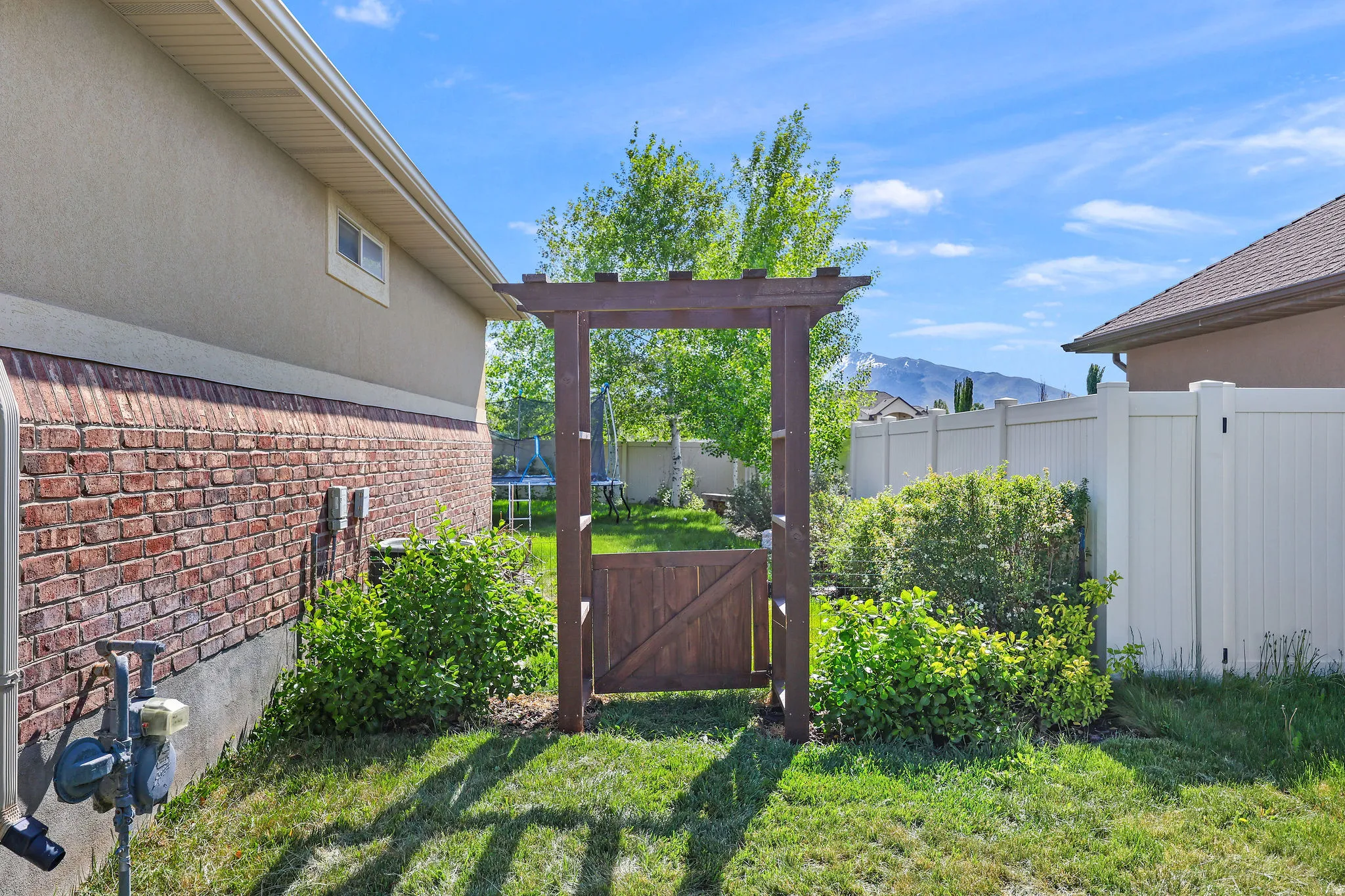 Landscaped backyard featuring a wooden arbor with a privacy gate, manicured lawn, established shrubs, and a neutral-toned vinyl fence