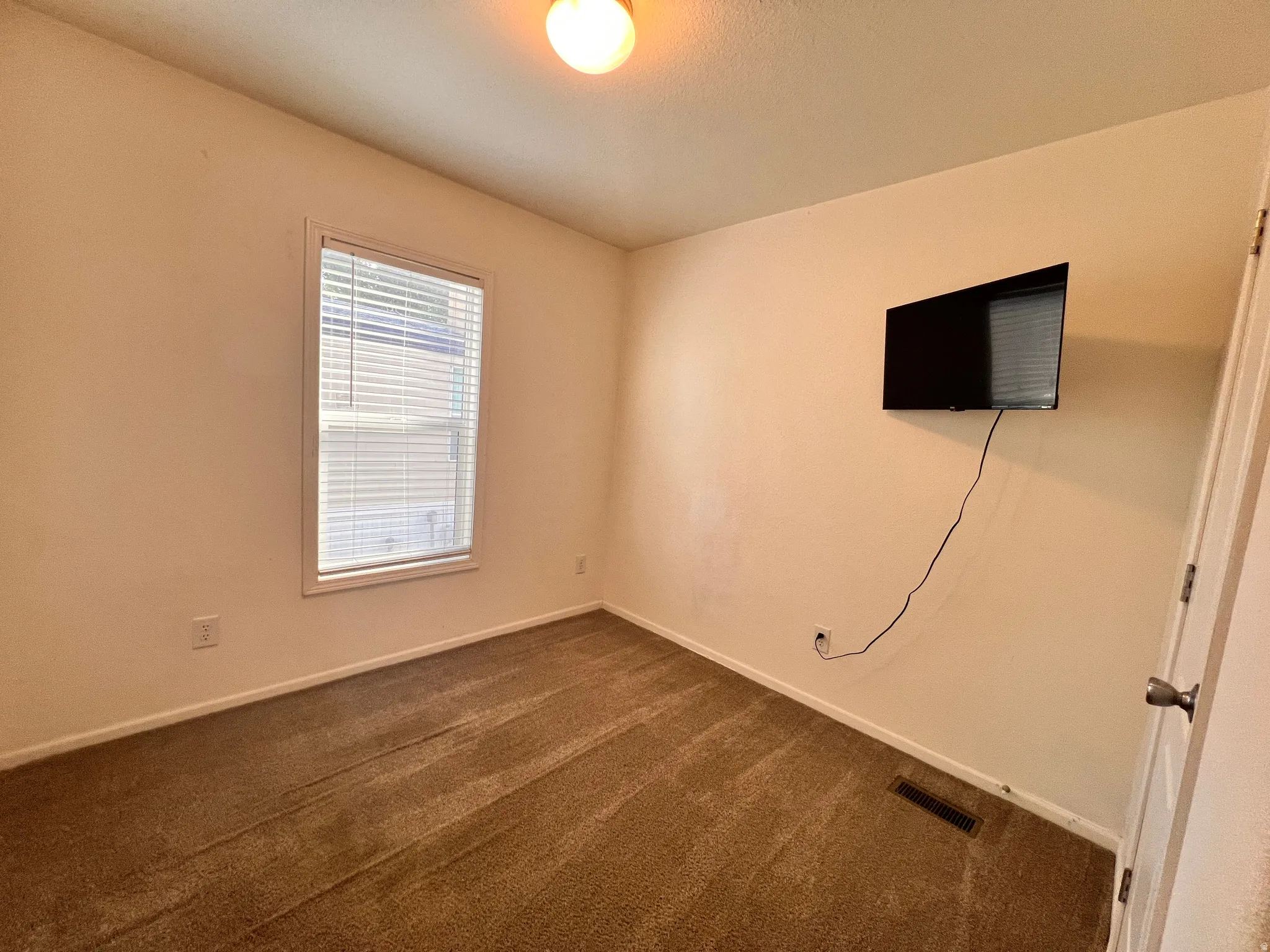 Carpeted room featuring neutral wall tones, a single window with blinds, and a ceiling-mounted light fixture. 32 inch tv included.