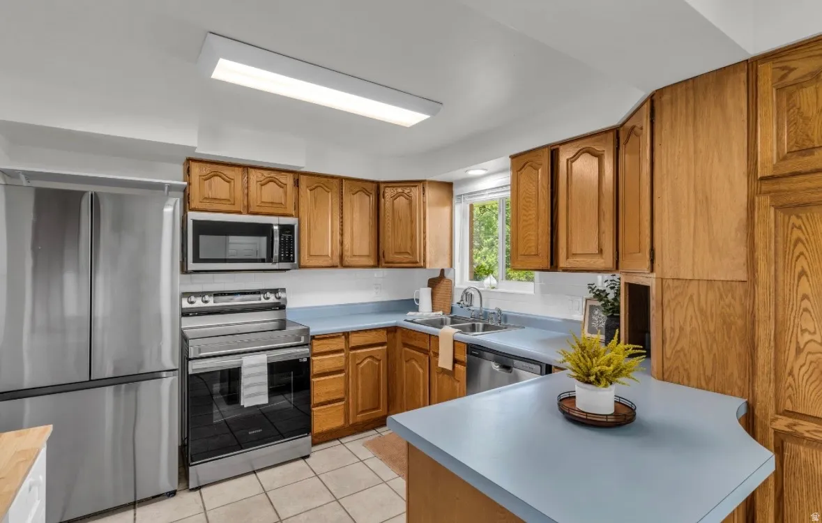 Kitchen featuring light wood cabinetry, light blue countertops, tile flooring, stainless steel appliances, and a window above the sink