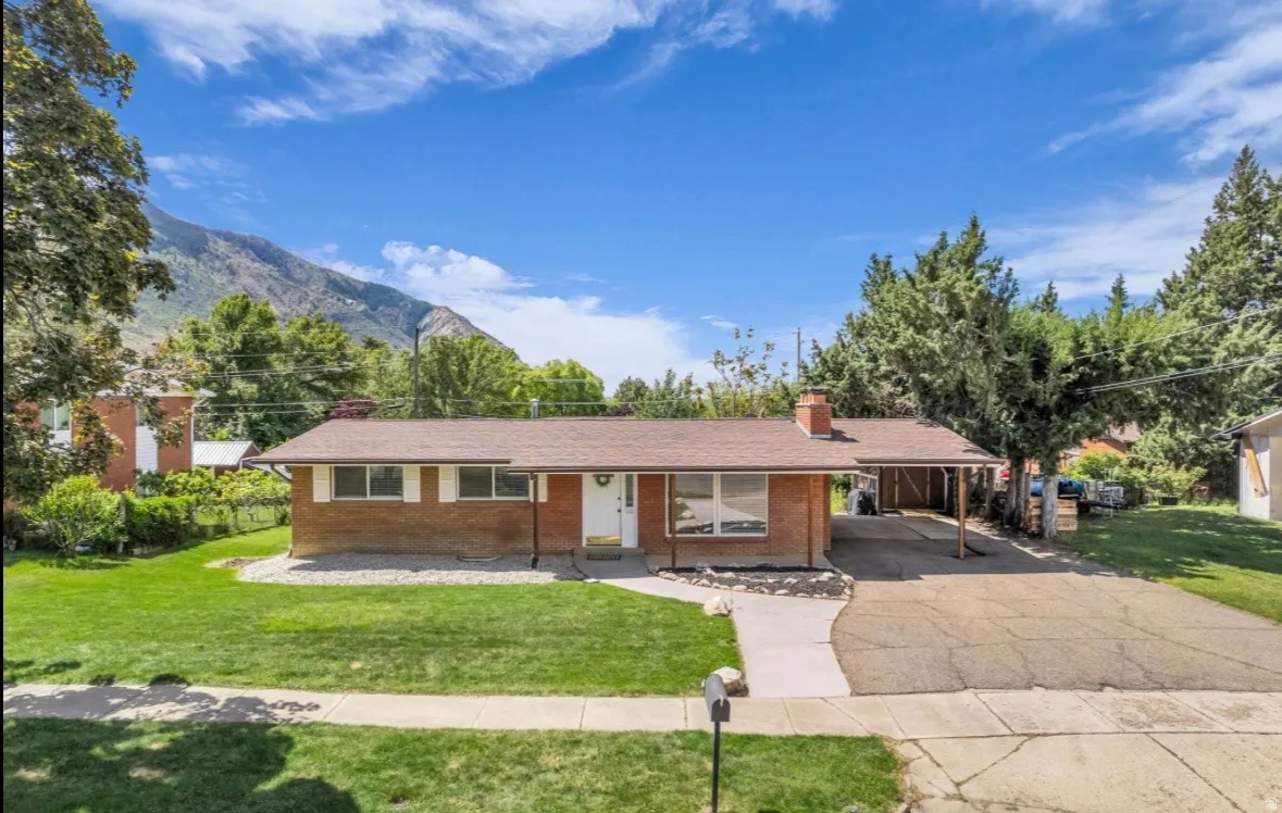 Brick exterior residence featuring a carport, front yard with mature landscaping, and an adjacent paved driveway
