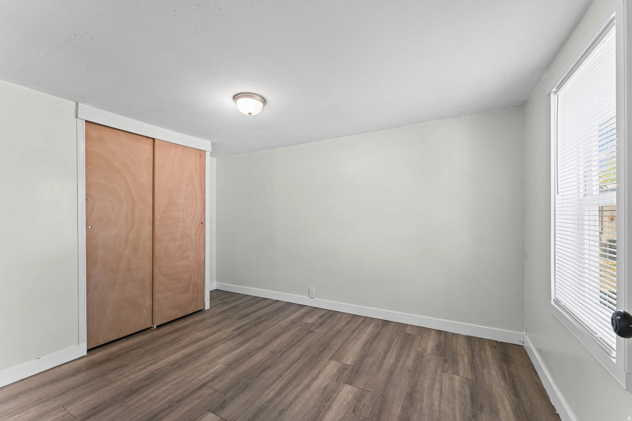 Room featuring wood-finish flooring, light-colored walls, a ceiling-mounted light fixture, a window with blinds, and a closet with wood sliding doors