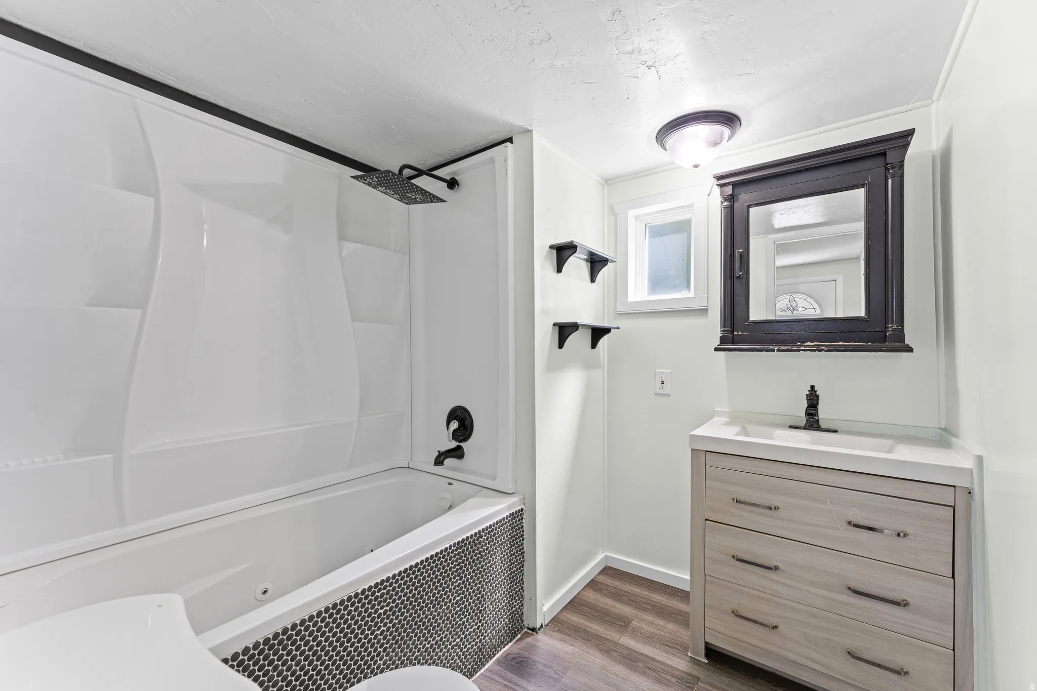 Bathroom featuring a white tub/shower combination with a black penny tile surround, a rain showerhead, a single vanity with light wood-finish drawers, a white countertop, and a dark wood-finish medicine cabinet