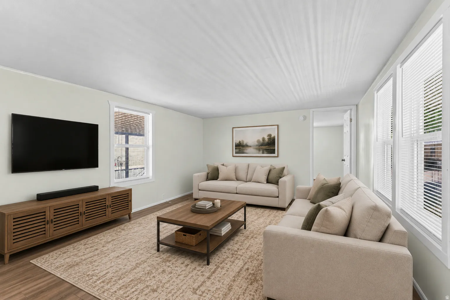 Spacious living area featuring light-toned walls, multiple windows with blinds, and wood-finish flooring