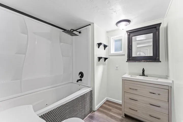 Bathroom featuring a white tub/shower combination with a black penny tile surround, a rain showerhead, a single vanity with light wood-finish drawers, a white countertop, and a dark wood-finish medicine cabinet