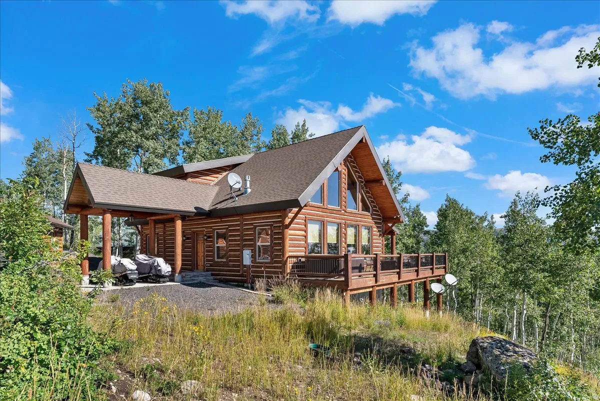 Log cabin exterior with an expansive wrap-around deck, large A-frame windows, covered entry, and a gravel driveway