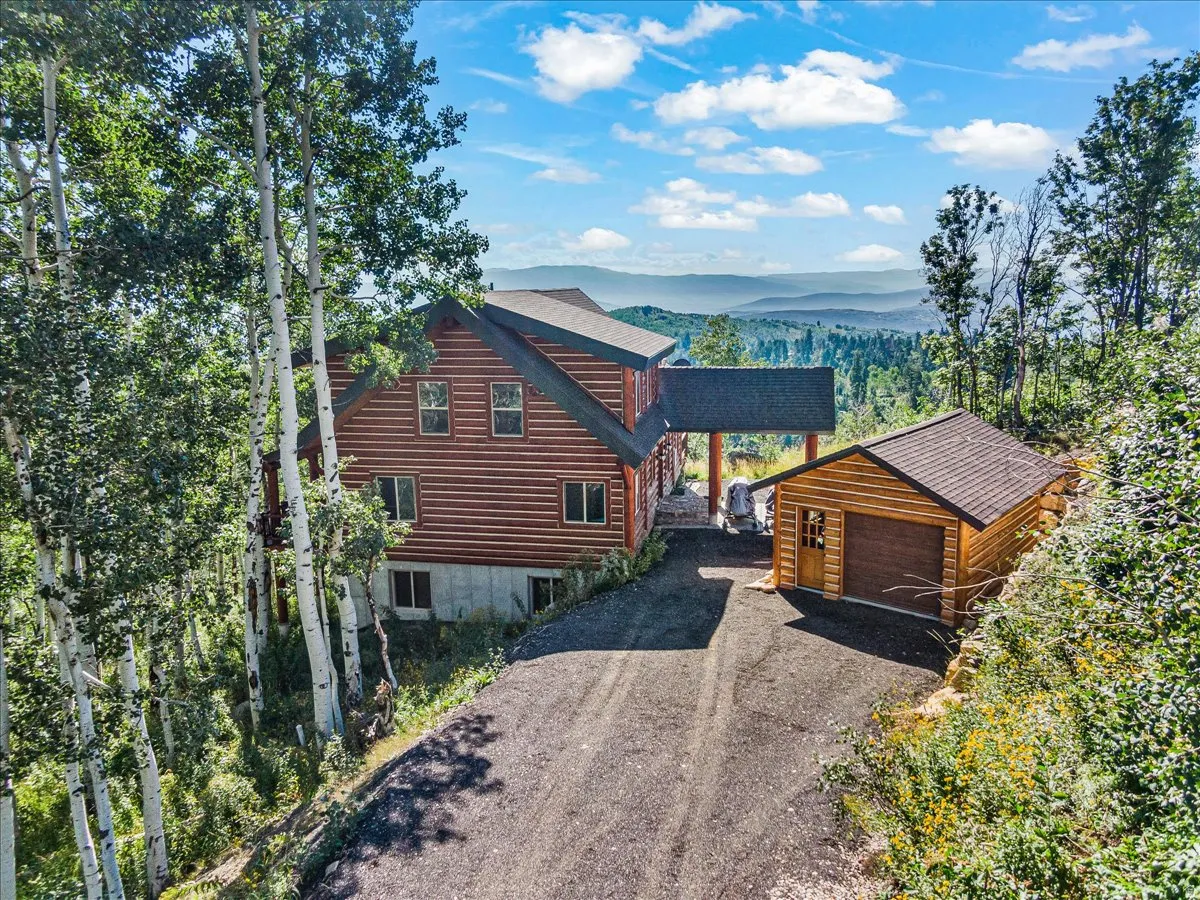 Log-style main residence with covered entry, separate garage structure, paved driveway, and mature trees on a sloped lot