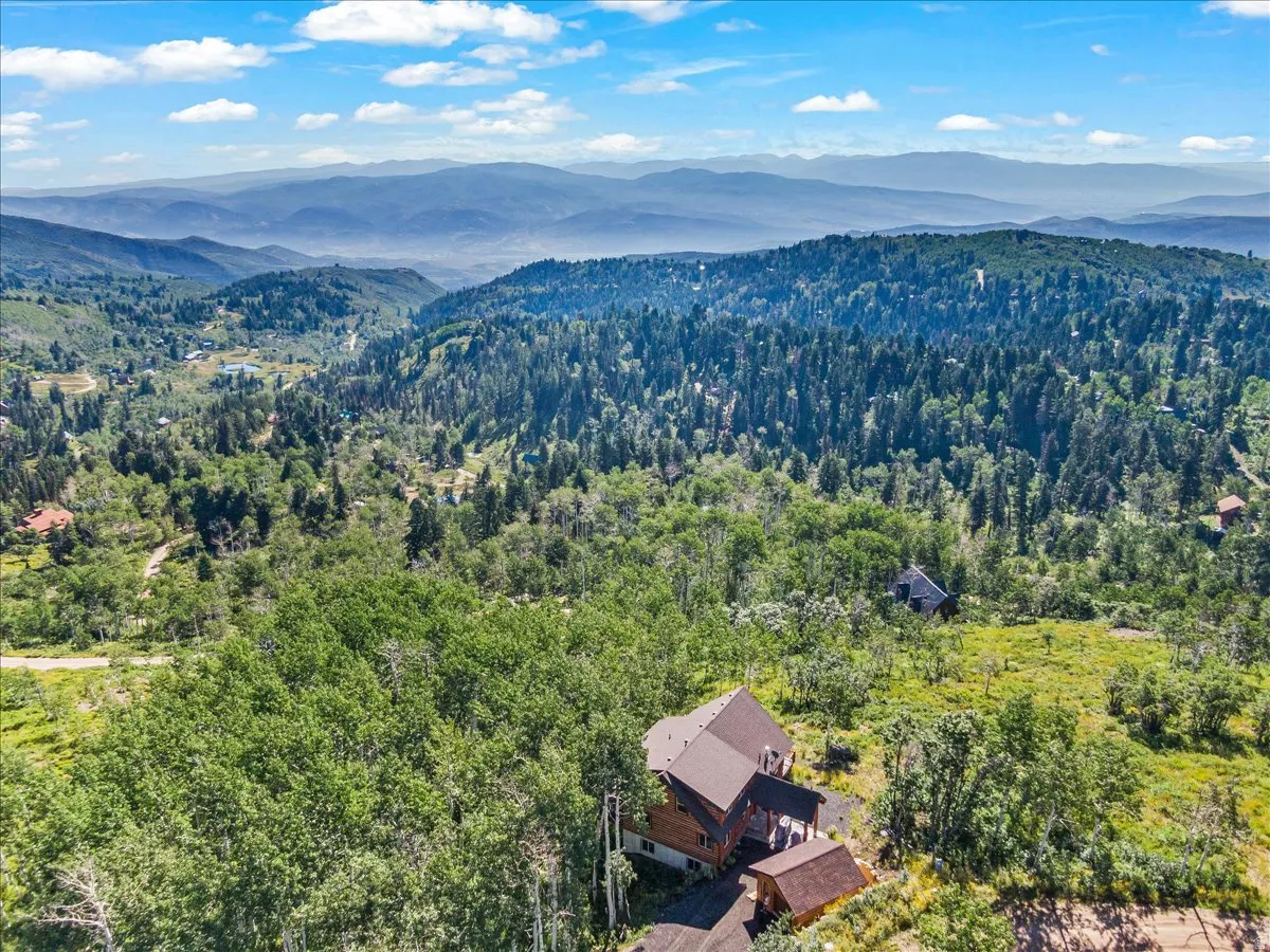 Expansive mountain landscape featuring a prominent residential structure with multiple levels and a brown roof, surrounded by dense green forestry