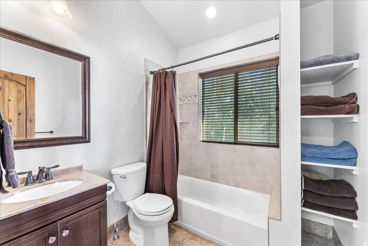 Bathroom featuring a tub/shower combination with tile surround, a built-in window with horizontal blinds, and a dark wood-finish vanity with a light-colored countertop and undermount sink