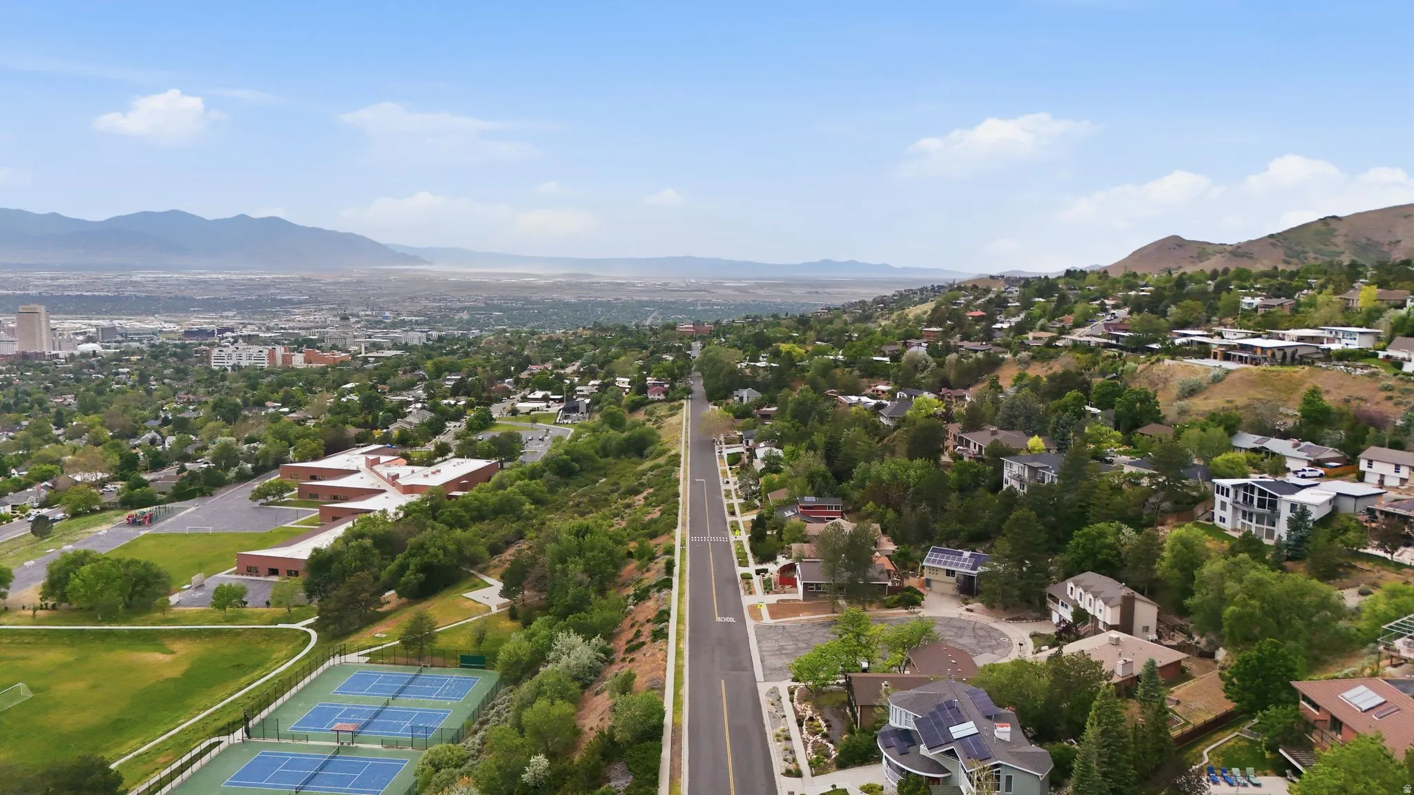 Expansive aerial view showcasing a hillside residential area with a prominent paved road, surrounded by mature trees