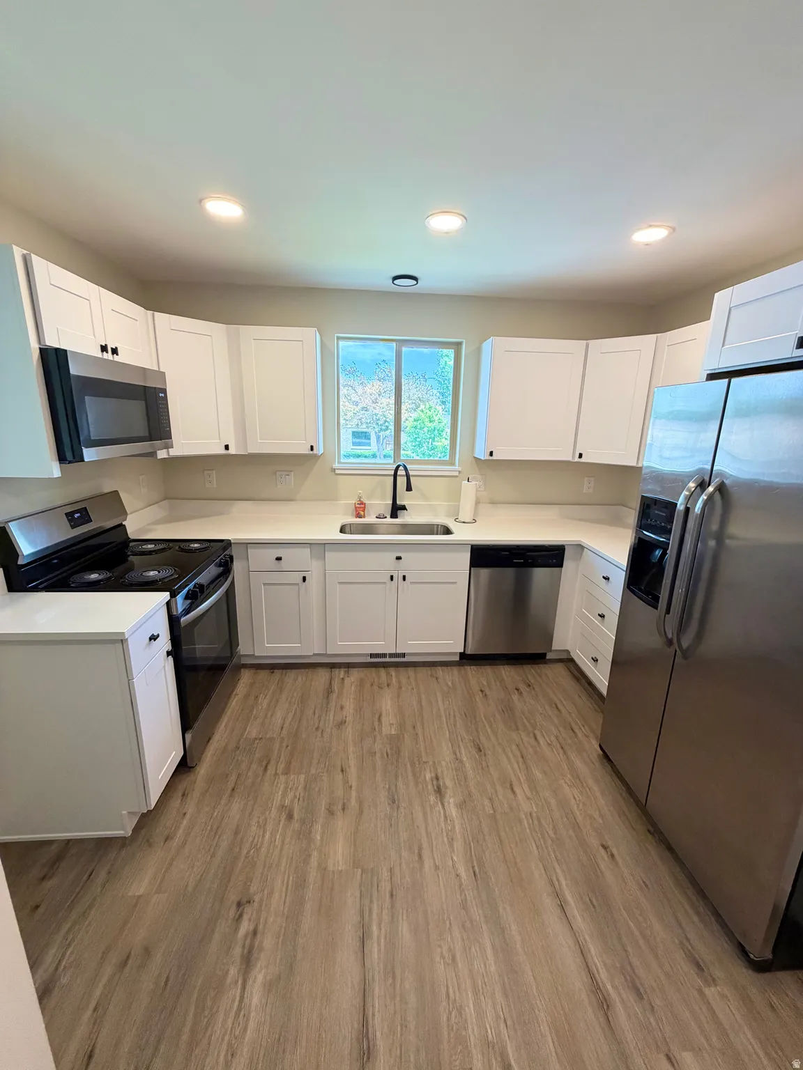 Modern kitchen featuring wood-finish flooring, white cabinetry, integrated stainless steel appliances, a window with exterior views, and recessed ceiling lighting