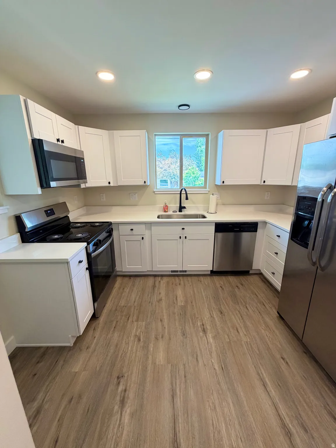 U-shaped kitchen featuring white shaker-style cabinetry, stainless steel appliances, light-toned countertops, wood-finish flooring, and recessed lighting
