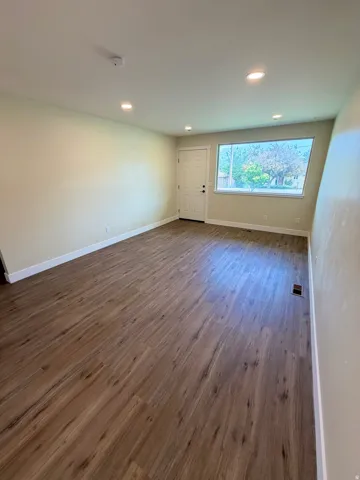 Spacious room featuring wood-finish flooring, recessed lighting, a white entry door, and a large front-facing window