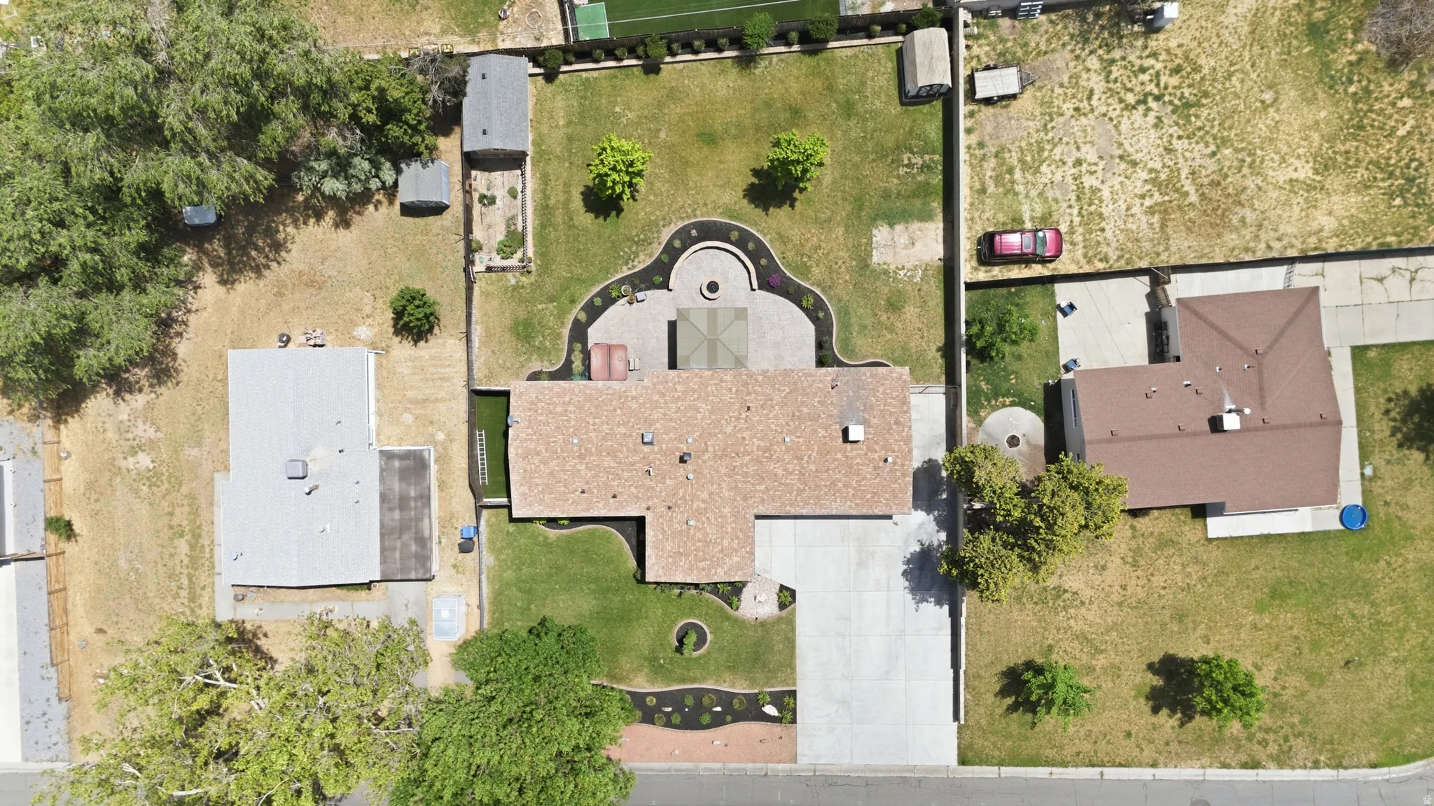 Aerial view highlighting a residential property with a brown shingle roof, a concrete driveway, a structured backyard patio, a grass lawn, and surrounding landscaping