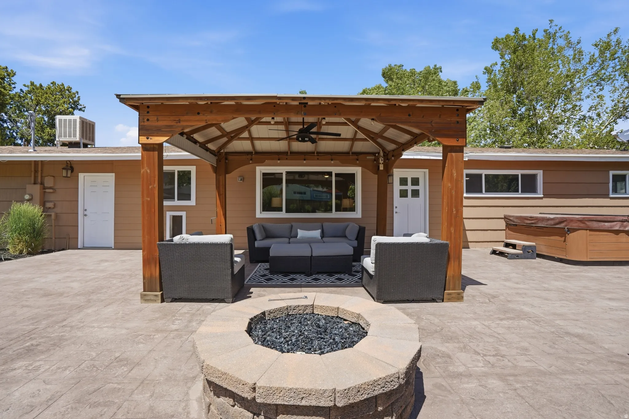 Expansive hardscaped patio featuring a substantial wood pergola with a ceiling fan, a built-in stone fire pit, and a separate hot tub