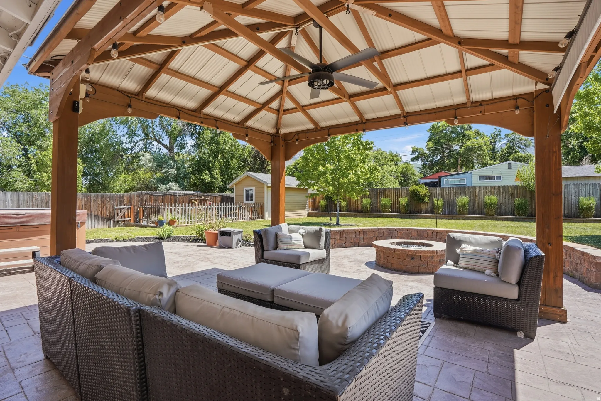 Expansive outdoor living area featuring a vaulted gazebo with exposed wood beams and a ceiling fan, a stamped concrete patio, a built-in circular fire pit with a stone surround, string lighting, and a fenced perimeter