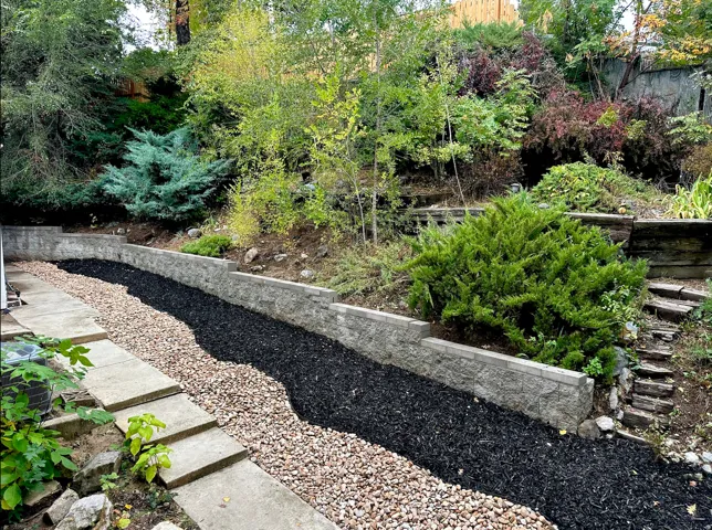 Tiered landscaping featuring concrete block retaining walls, organic shaped beds with dark mulch and light gravel, and mature shrubbery