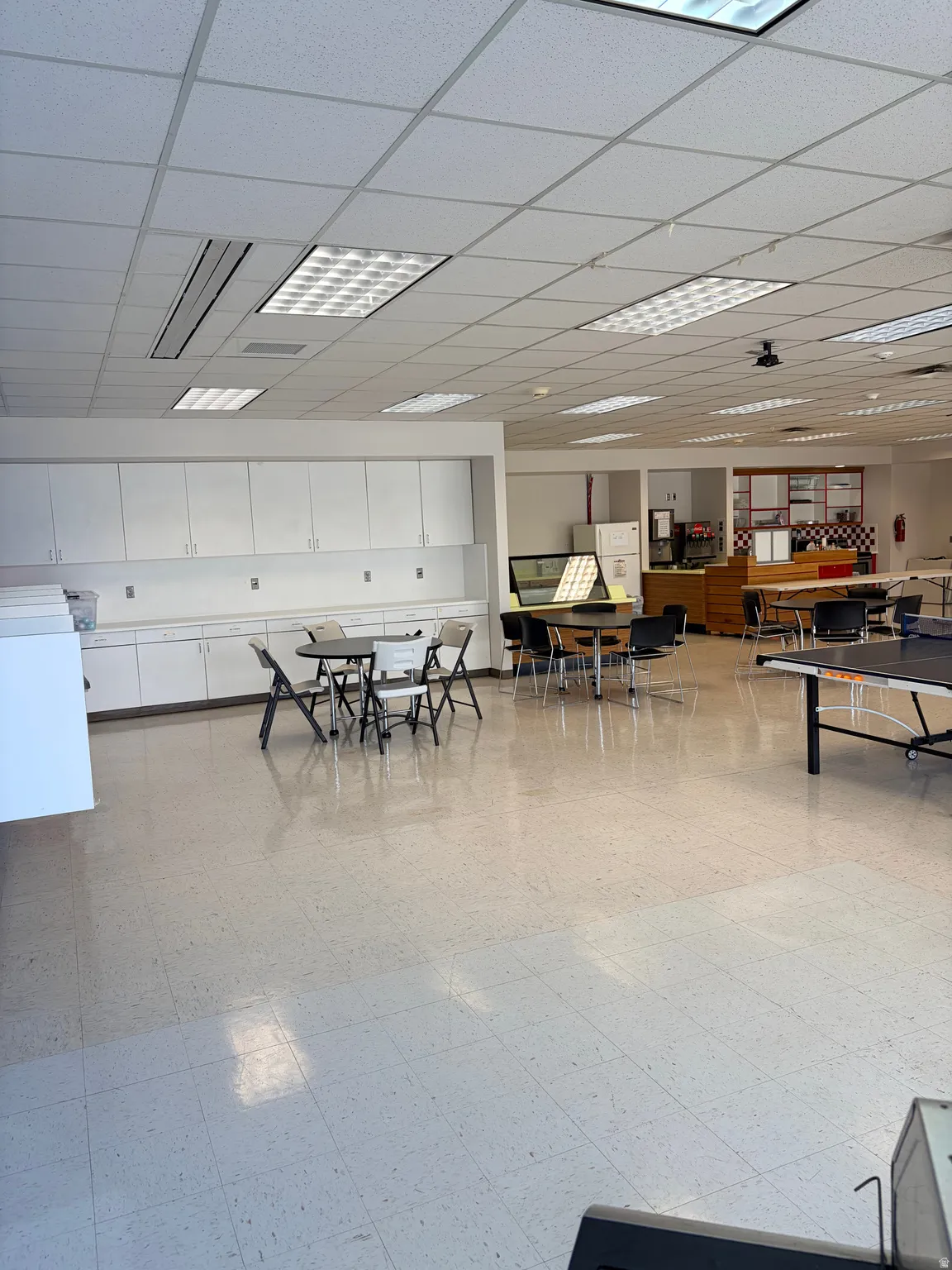 Spacious interior featuring a grid ceiling with integrated fluorescent lighting, extensive built-in white cabinetry, a light-toned tile floor, a built-in counter space, and a serving area with display shelving