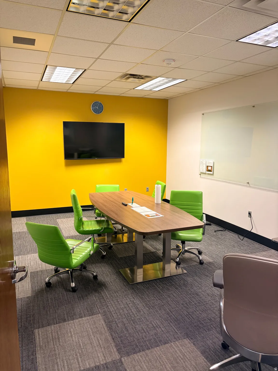 Conference room featuring a wall-mounted display, wall-mounted whiteboard, wood-finish conference table with metal bases, and modular carpet flooring