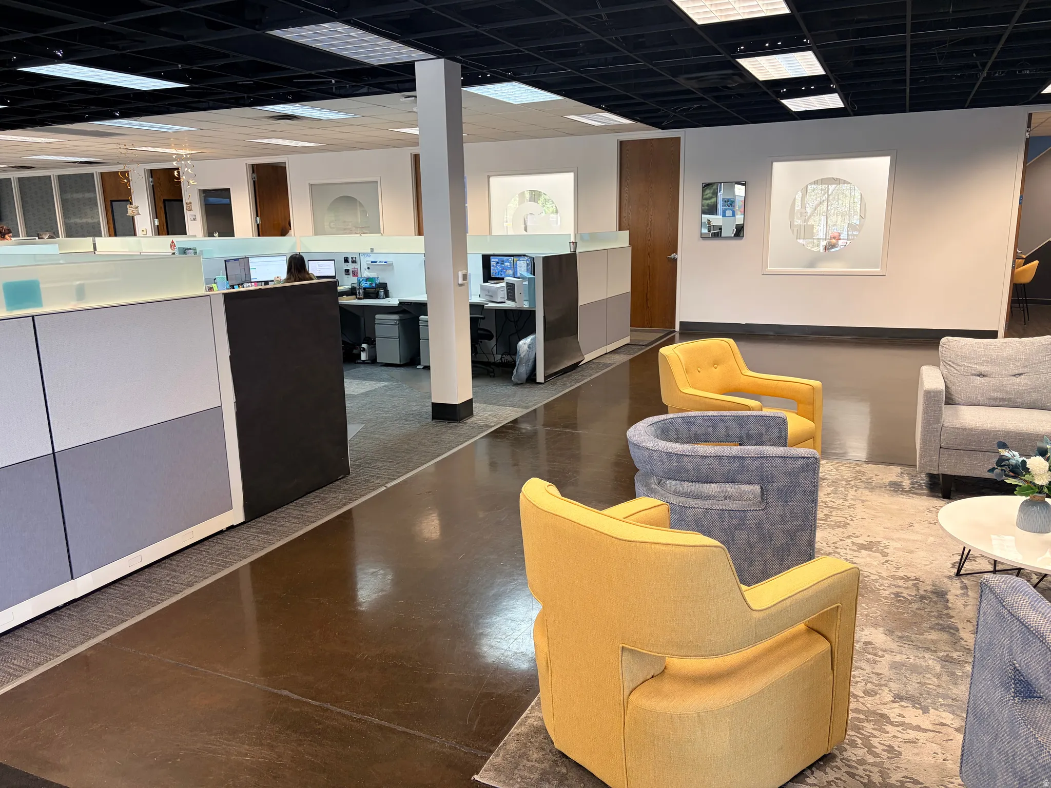 Open-plan office space featuring a mixed flooring of polished concrete and carpeting, a black grid ceiling with recessed lighting, and a lounge area