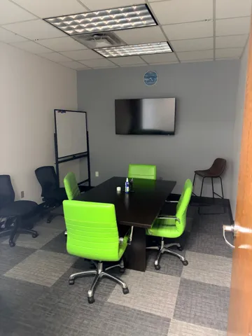 Conference room featuring a wall-mounted display, grid ceiling with recessed lighting, and checkerboard carpet tile flooring