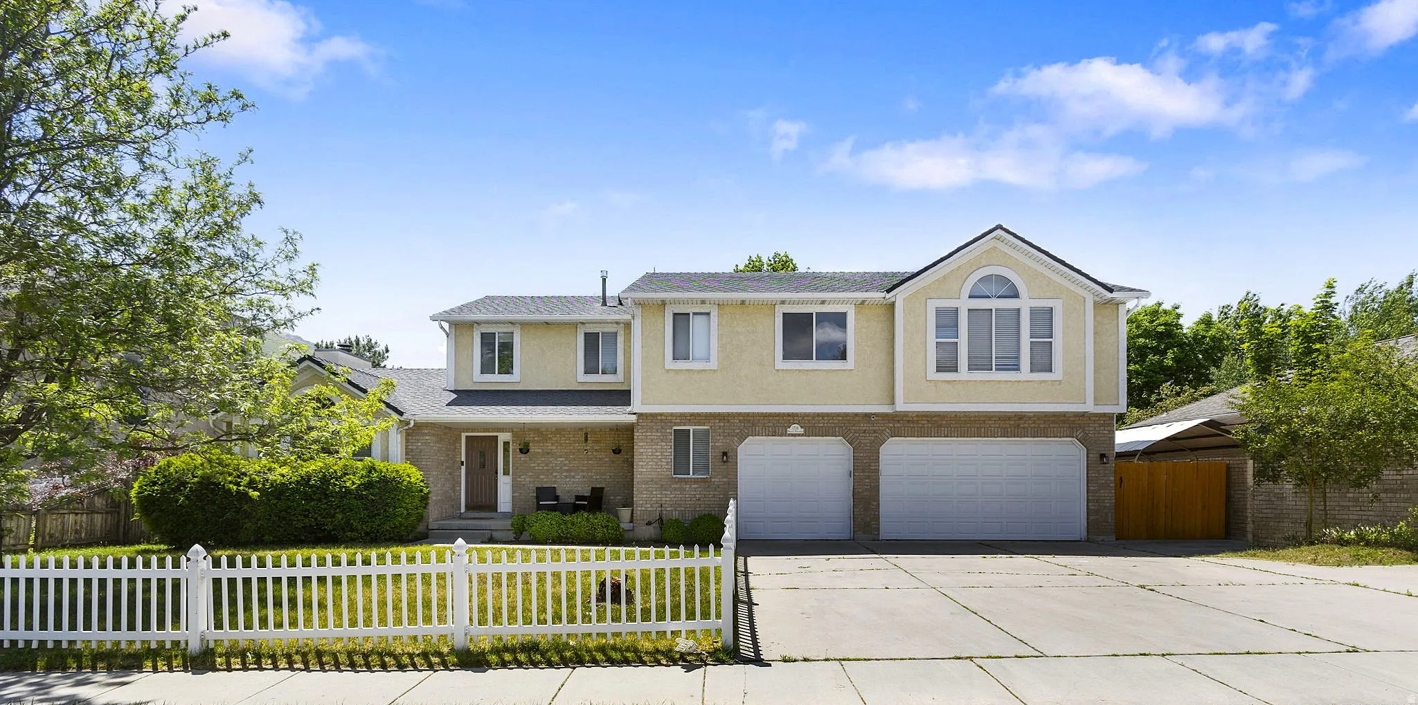 Two-story residence featuring a stucco and brick facade, dual garage doors, a white picket fence, a covered porch, and an arched window architectural detail