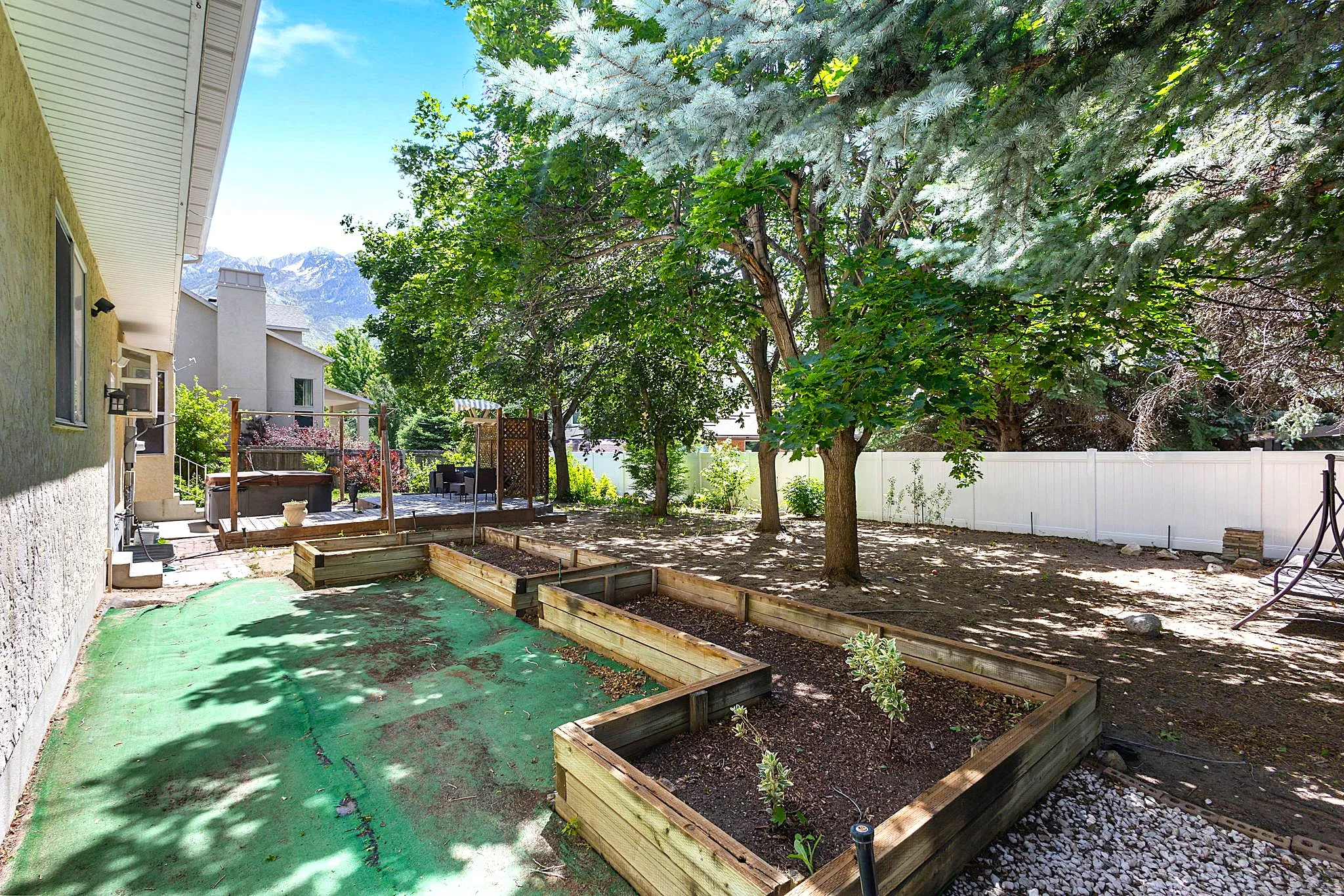 Spacious backyard featuring multiple raised garden beds, mature trees, a white perimeter fence, a built-in deck, and mountain scenery