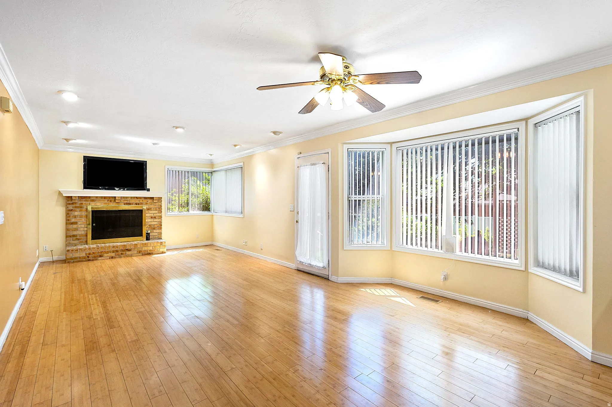Spacious room featuring wood-finish flooring, a brick-surround fireplace with a white mantel, and a multi-pane bay window with vertical blinds
