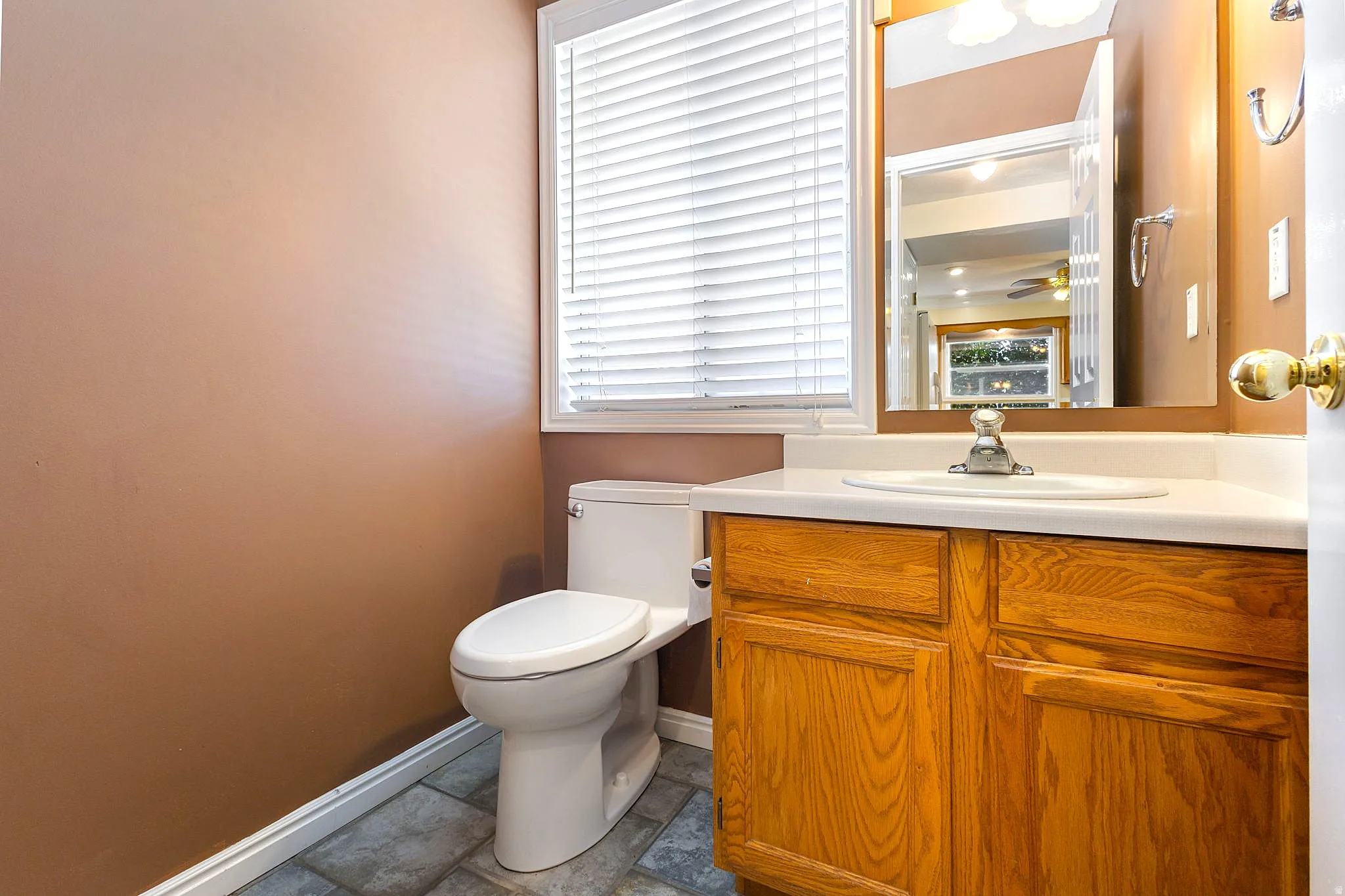 Bathroom featuring a wood-finish vanity with an integrated sink, a wall-mounted mirror, a toilet, a window with blinds, and tile flooring