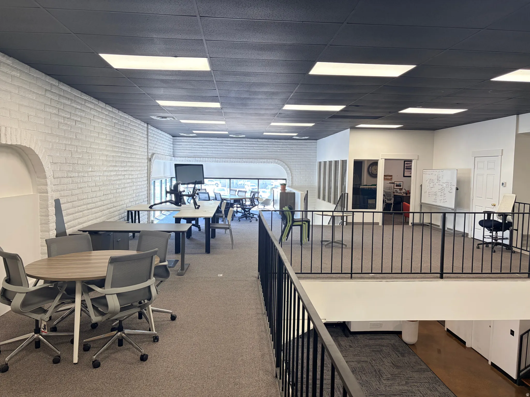 Open-concept office space featuring carpeted flooring, white painted brick walls, and a black grid ceiling with recessed lighting