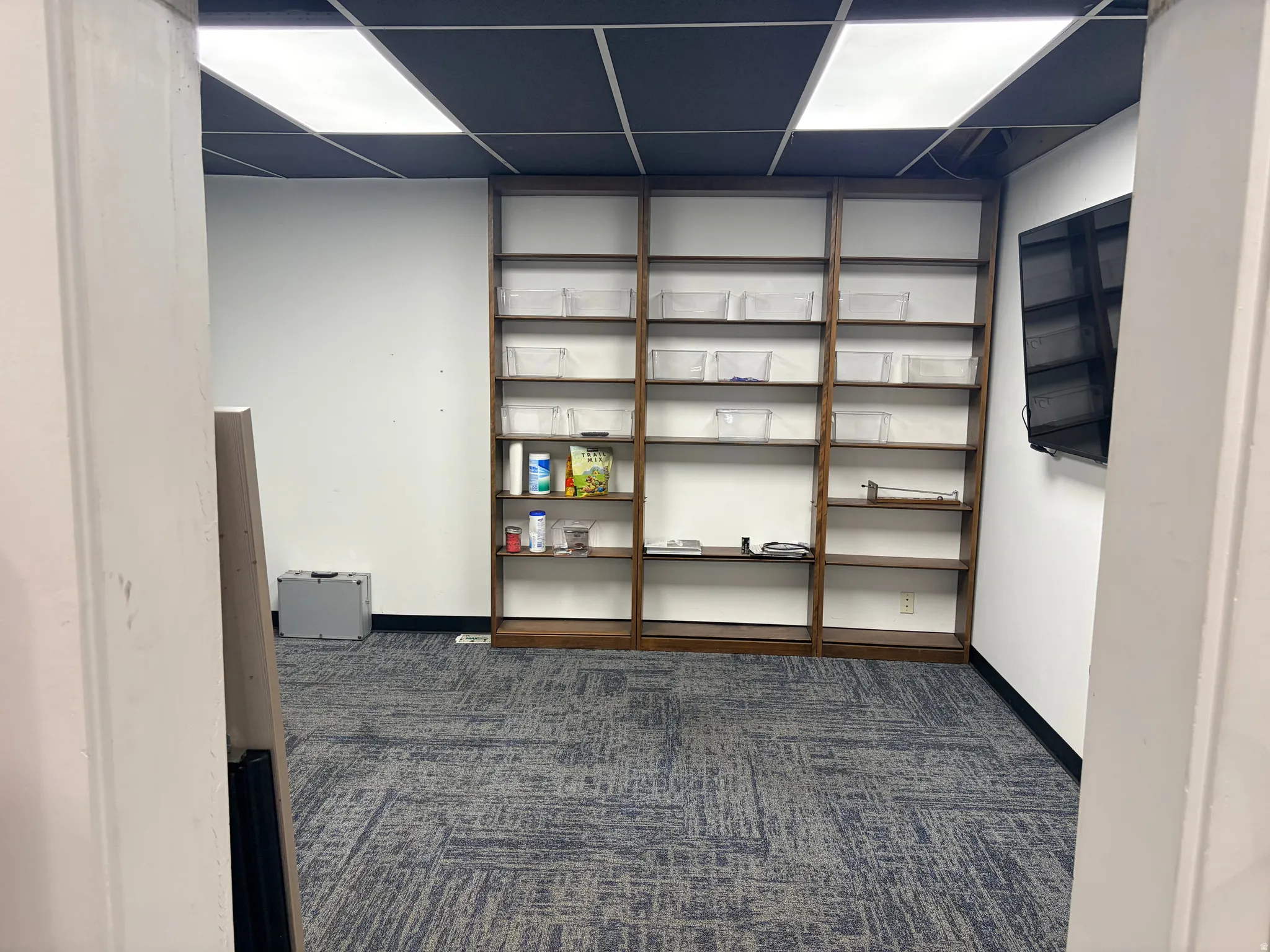 Room featuring dark wood-finish built-in shelving, patterned carpet flooring, and a suspended ceiling with integrated lighting panels