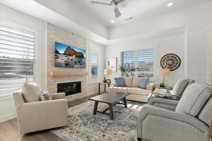 Living area featuring a textured stone fireplace surround, wood-finish flooring, plantation shutters, a coffered ceiling, and a ceiling fan with integrated lighting