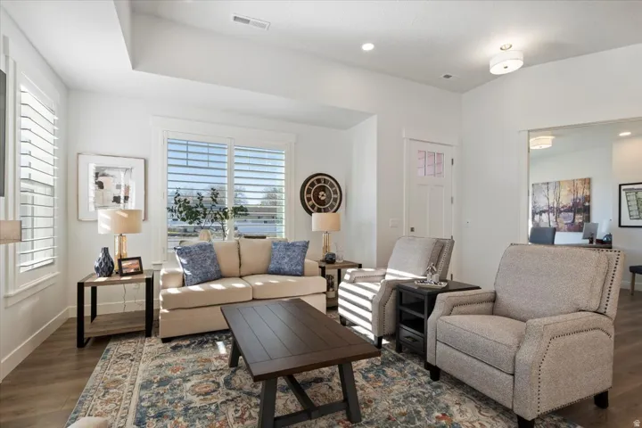 Bright living area featuring wood-finish flooring, white plantation shutters, tray ceiling, and a paneled entry door with frosted glass