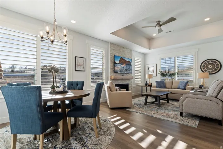 Open-concept living space featuring wood-finish flooring, a stone-clad fireplace with a wood mantle, a tray ceiling with a ceiling fan, recessed lighting, and multiple windows with plantation shutters
