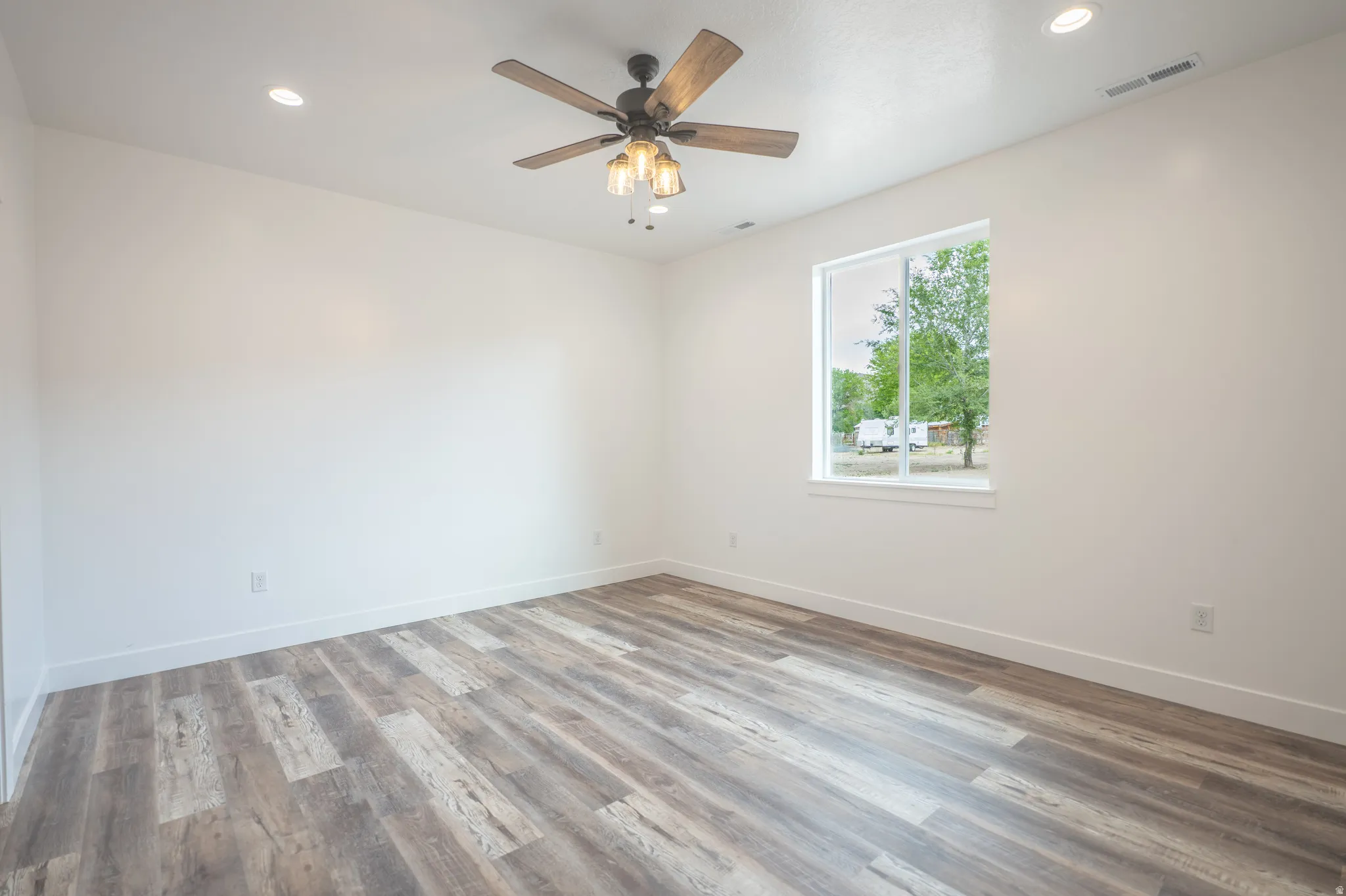 Spacious room featuring wood-finish flooring, a ceiling fan with light fixture, recessed lighting, and a window with white trim