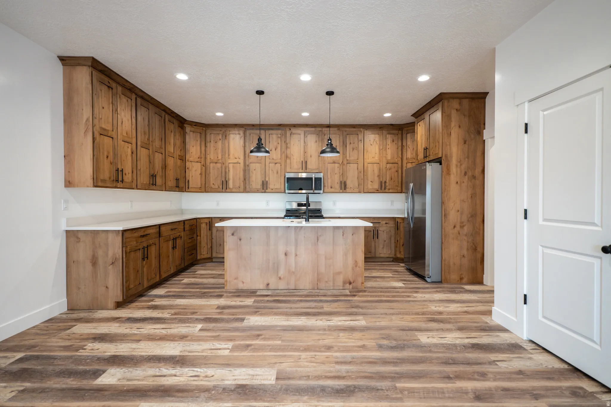 Open-concept kitchen featuring extensive wood cabinetry, white countertops, a central island with a built-in cooktop, stainless steel appliances, and wood-finish flooring