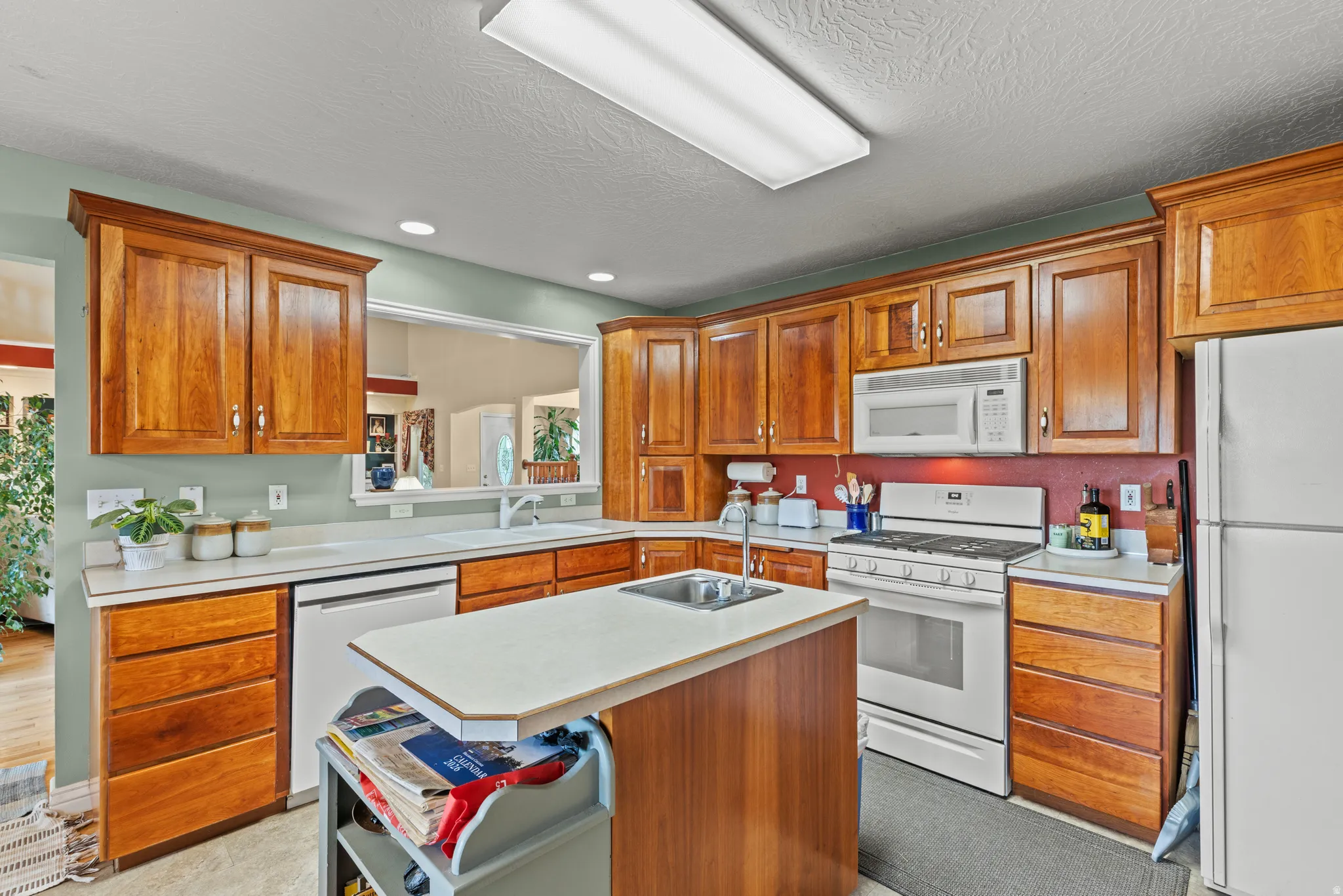 Kitchen featuring wood cabinetry, white countertops, a central island with an integrated sink, white appliances, and a pass-through window