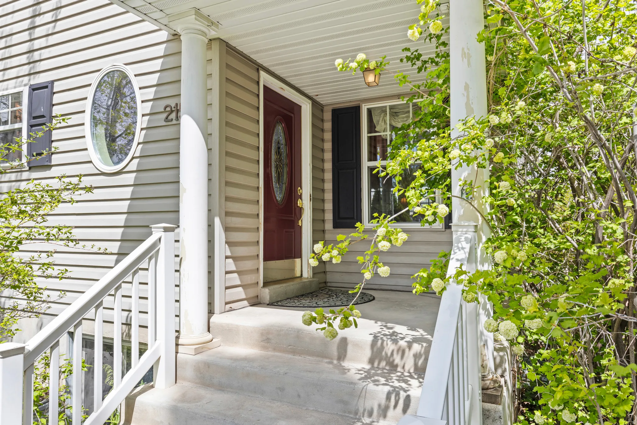 Inviting front entry featuring a red paneled door with oval glass insert, a complementary oval window, light-toned siding, and a covered porch with white columns and railings
