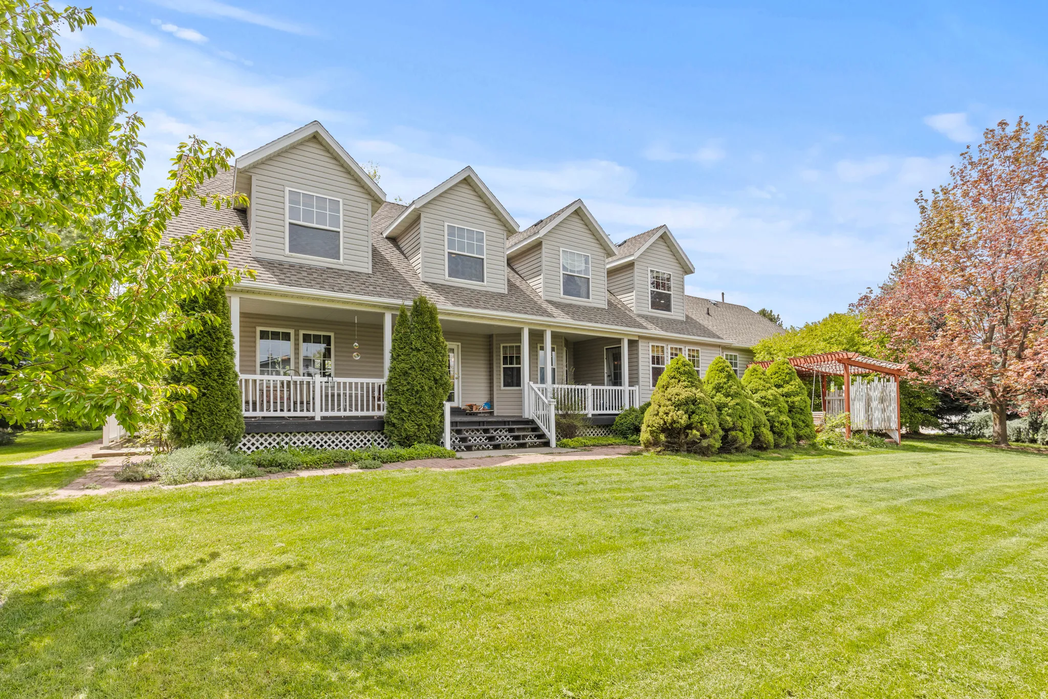 Exterior featuring light gray siding, multiple dormers, an expansive front lawn, a covered porch with white railings, and a wooden pergola