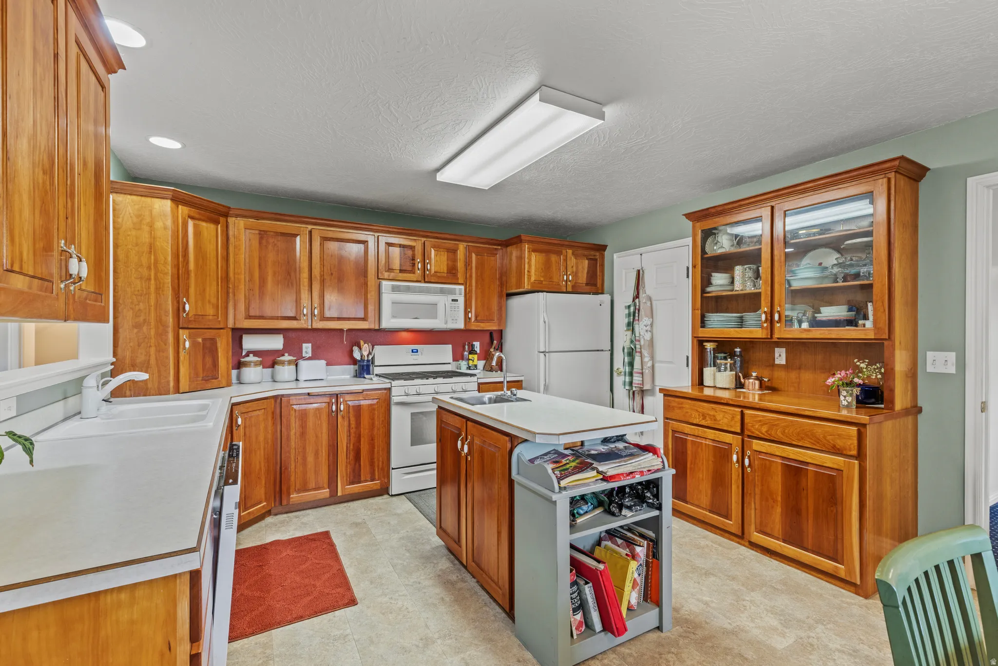 Kitchen featuring extensive wood cabinetry, a central island with shelving, white appliances, recessed lighting, and a built-in hutch with glass-front display cabinets