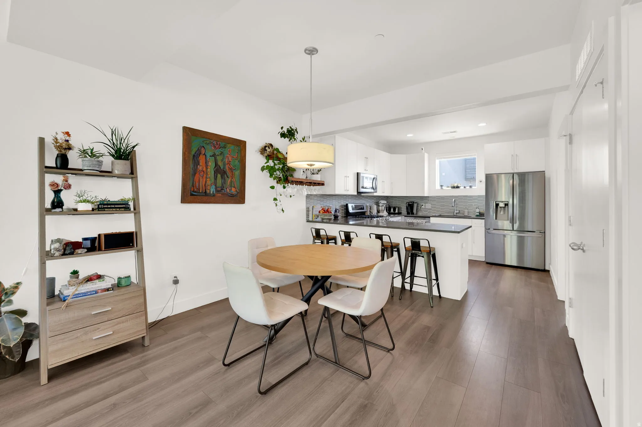 Open-concept living space featuring wood-finish flooring, a contemporary kitchen with stainless steel appliances, white cabinetry, dark countertops, and a tiled backsplash
