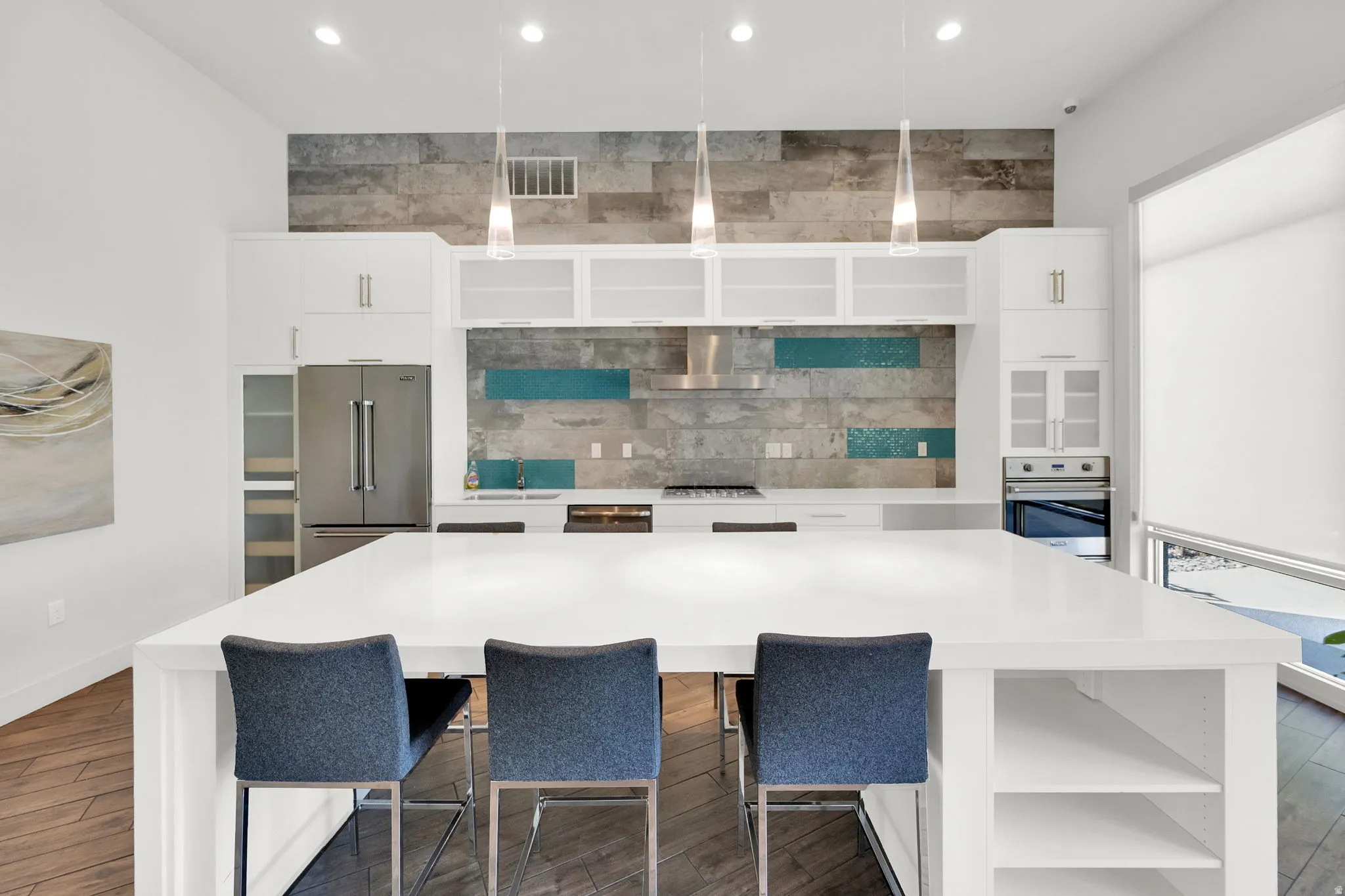 Spacious kitchen featuring a large white island with integrated shelving, stainless steel appliances, a unique tile backsplash with blue accents, white cabinetry, and pendant lighting
