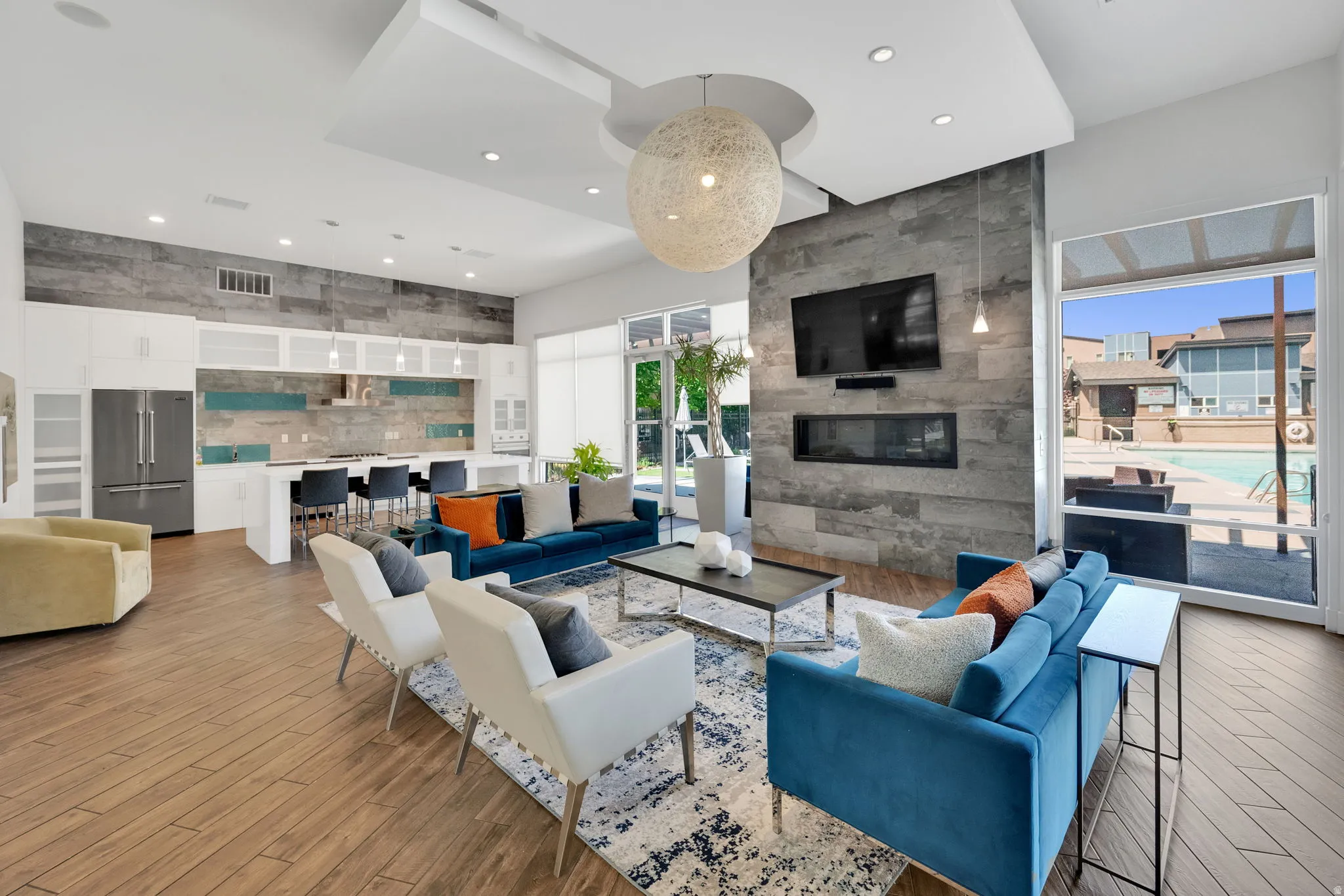 Open-concept living space with wood-finish flooring, a contemporary fireplace feature wall, a full kitchen with stainless steel appliances, and large windows overlooking a swimming pool
