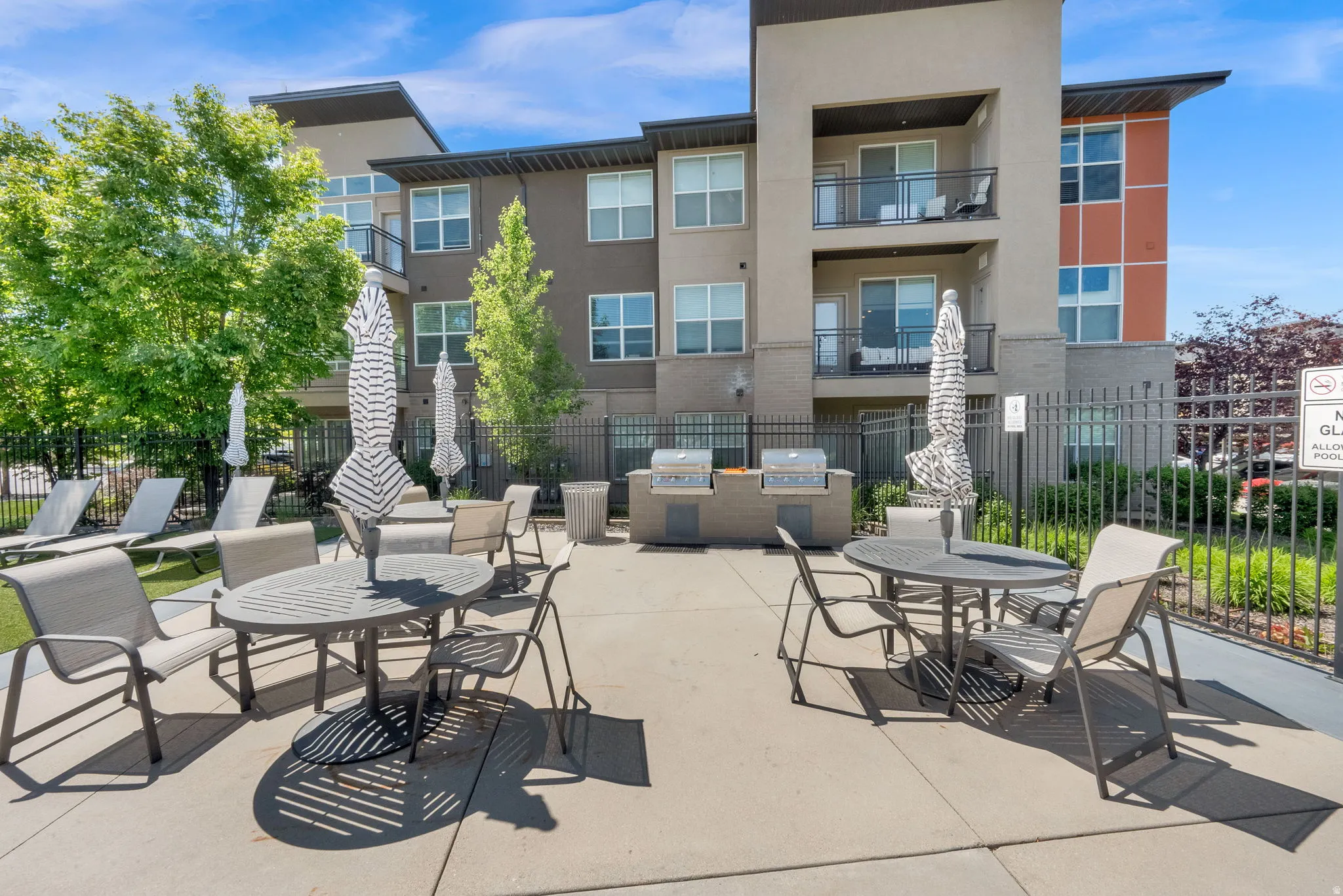 Outdoor patio area featuring multiple dining tables with umbrellas, lounge seating, a barbecue station with two grills, and a black metal fence