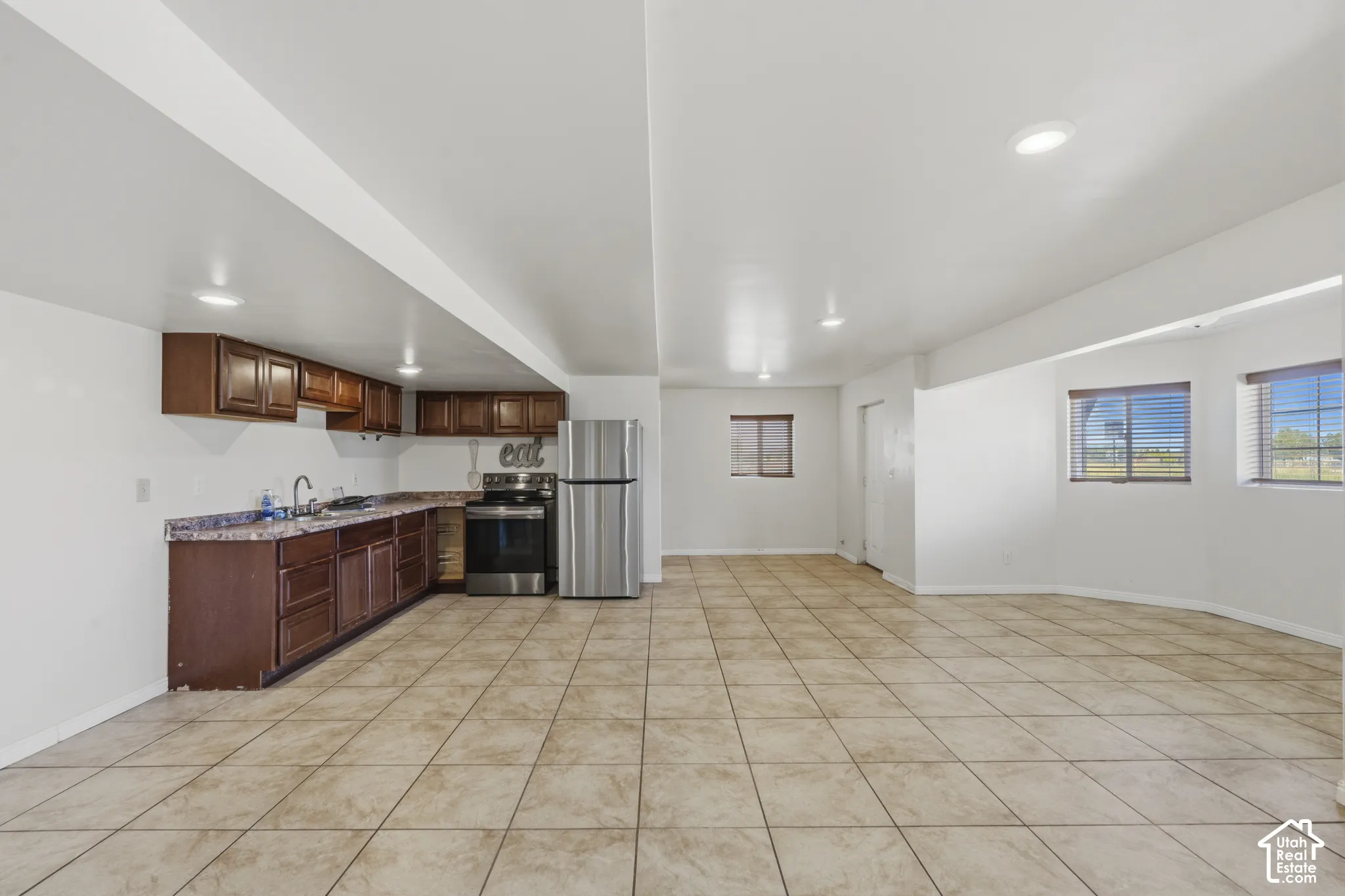 Expansive open-concept room featuring a functional kitchen area, stainless steel appliances, dark wood-finish cabinetry, and neutral-toned tile flooring