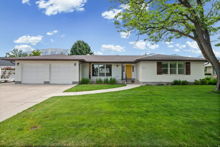 Ranch-style exterior featuring white brick, an expansive front lawn, and a light-colored driveway leading to a two-car garage