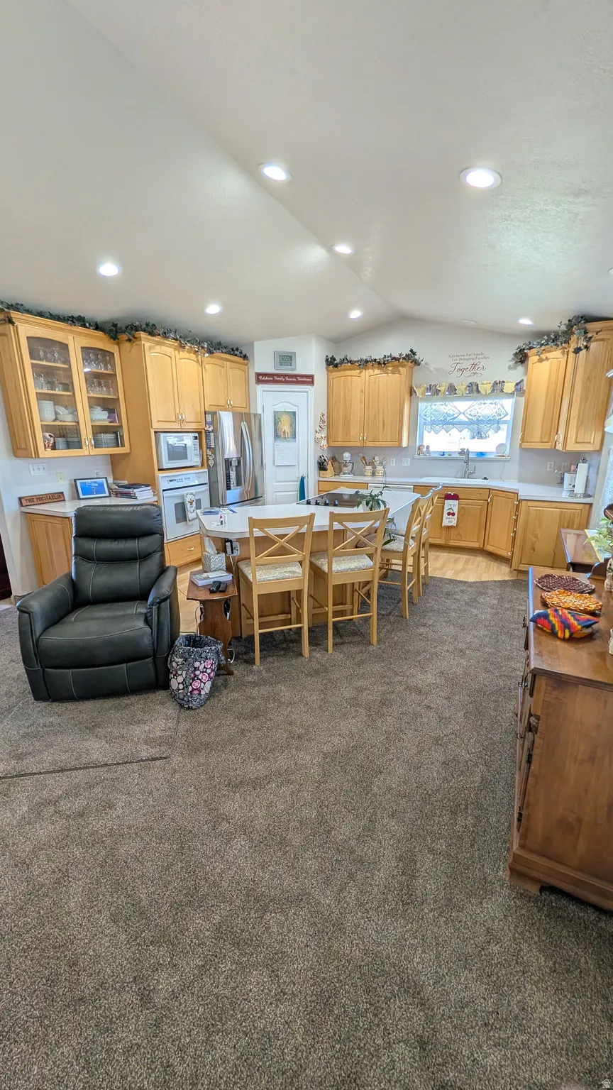 Open-concept kitchen with light wood cabinetry, recessed lighting, a central island with seating, a stainless steel refrigerator, and a double-basin sink under a window