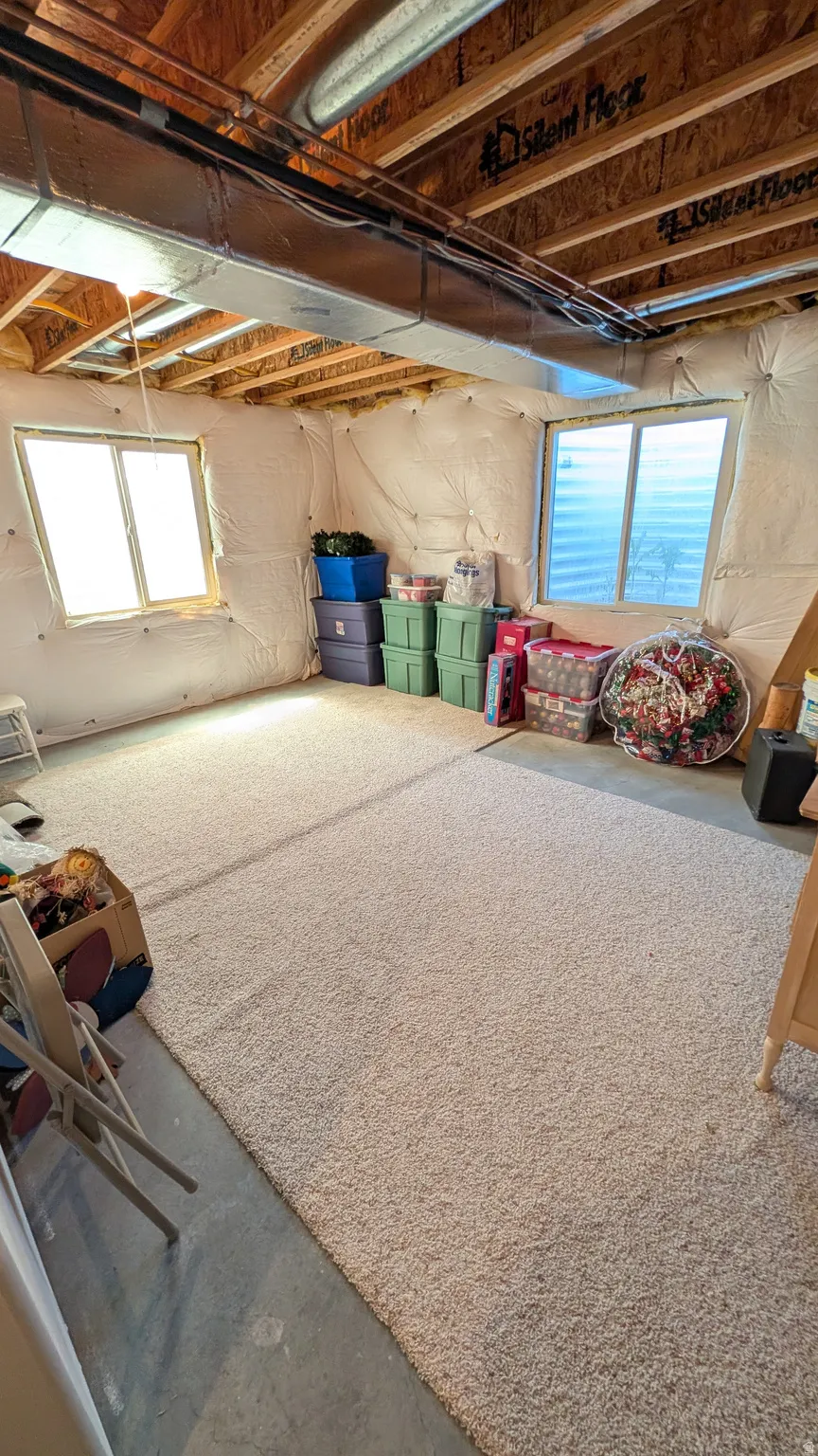 Spacious unfinished basement offering two windows, exposed ceiling joists, ductwork, and a light-colored carpeted area
